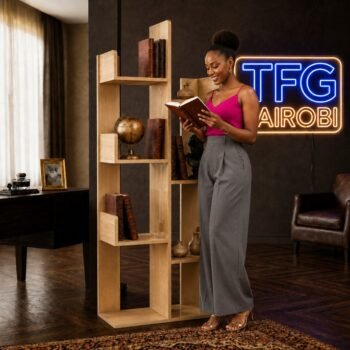 A smiling woman in a pink top and grey trousers reading a book beside a TFG TR600-5 natural oak tree bookshelf in a luxury home office on Mombasa Road.