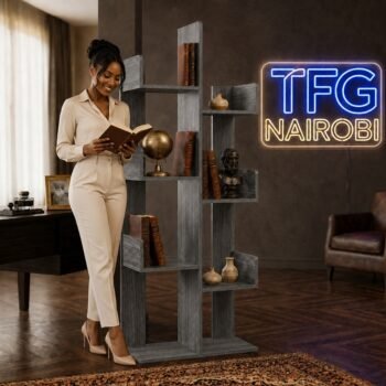 A smiling woman in an elegant beige jumpsuit standing beside the TFG TR600-3 storm grey tree bookshelf and reading a book in a luxury home office on Amboseli Road.